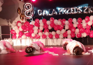 Dziewczynki w białych sukienkach są na scenie w siadzie skulnym. Są również balony i napis Gala Przedszkolaka.